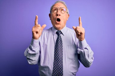 Gri saçlı, gözlüklü kıdemli iş adamı mor izole edilmiş arka plan üzerinde duruyordu. Şaşırmış ve şaşırmıştı. Parmaklarıyla işaret ediyor ve kollarını kaldırıyordu..