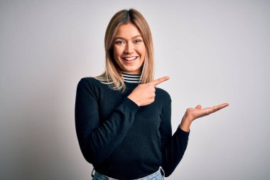 Genç, güzel, mavi gözlü, beyaz arka planda duran sarışın kadın hayrete düşmüş ve kameraya gülümsüyor, eliyle ve parmağıyla işaret ediyor..