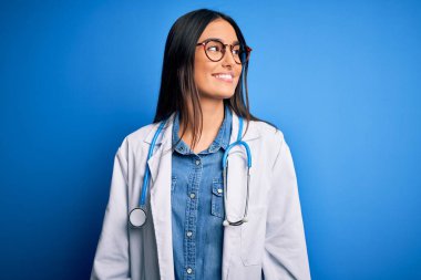 Steteskop takan ve mavi arka planda gözlük takan genç, güzel bir doktor. Yüzünde gülümseme, doğal bir ifade var. Kendinden emin gülen.