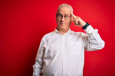 Orta yaşlı yakışıklı, beyaz gömlekli ve kırmızı arka planda gözlüklü bir adam. Tek parmakla kafasını göstererek gülümsüyor. Harika bir fikir ya da düşünce, iyi bir hafıza.