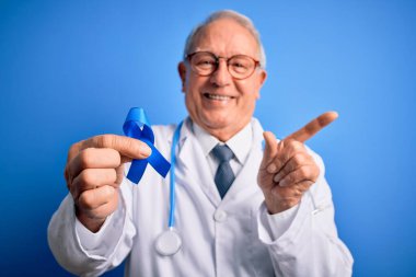 Kır saçlı kıdemli doktor elinde kolon kanseri farkındalığı mavi arka planda mavi kurdele çok mutlu bir şekilde el ve parmak kenarı gösteriyor.