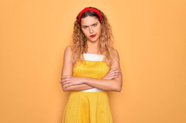 Sarı arka plan üzerinde dikey duran mavi gözlü güzel sarışın kadın şüpheci ve gergin, yüzünde çapraz kollu tasvip etmeyen ifade. Negatif kişi.