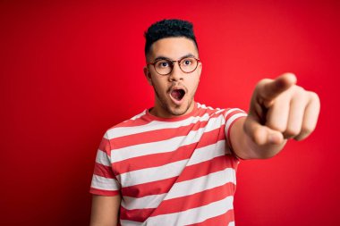 Genç ve yakışıklı bir adam gündelik çizgili tişört ve gözlük takıyor. Kırmızı arka plan üzerinde. Parmağıyla işaret ediyor.