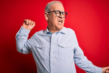 Orta yaşlı, yakışıklı, beyaz gömlekli, kırmızı arka planda gözlüklü mutlu ve neşeli dans eden, hareketli, rahat ve kendinden emin müzik dinleyen bir adam.