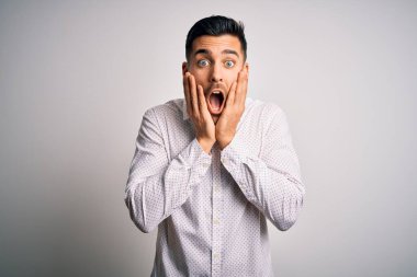 Zarif bir gömlek giyen yakışıklı genç adam izole edilmiş beyaz arka planda duruyor. Korkmuş ve şok olmuş, şaşırmış ve şaşırmış bir yüz ifadesi var.