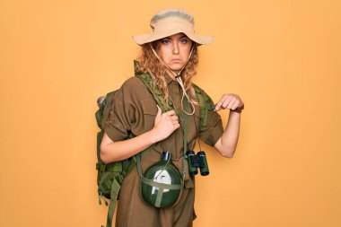 Mavi gözlü genç sarışın kaşif sırt çantası ve su kantini takıyor üzgün ve üzgün görünüyor, parmaklarıyla yön gösteriyor, mutsuz ve depresif..