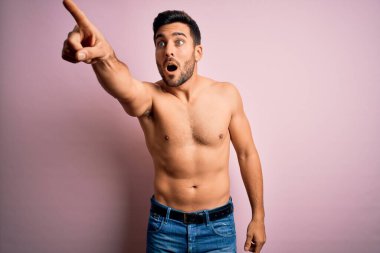 Sakalsız, genç, yakışıklı, güçlü bir adam pembe arka planda dikiliyor. Parmakları önde, ağzı açık, şaşırmış bir şekilde işaret ediyor.