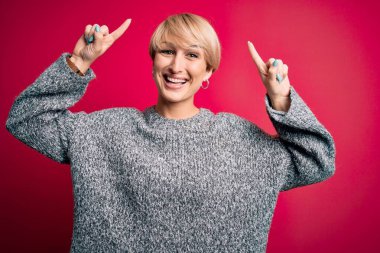 Modern kısa saçlı genç bir kadın pembe arka planda rahat bir süveter giyiyor. Şaşırmış, şaşırmış ve parmaklarıyla işaret ediyor ve kollarını kaldırıyor..