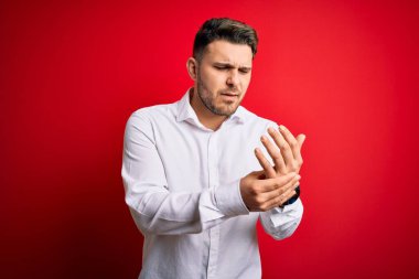 Mavi gözlü, zarif gömlekli genç bir iş adamı kırmızı izole edilmiş arka planda duruyor. Ellerinde ve parmaklarında ağrı var. Eklem iltihabı.