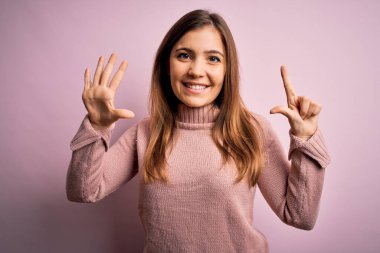 Güzel genç bir kadın pembe izole edilmiş arka planda balıkçı yaka kazak giyiyor ve yedinci parmaklarıyla işaret ederken kendinden emin ve mutlu gülümsüyor..