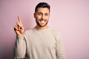 İzole edilmiş pembe arka plan üzerinde duran sıradan süveter giyen genç ve yakışıklı bir numaralı parmağını göstererek kendinden emin ve mutlu bir şekilde gülümsüyor..