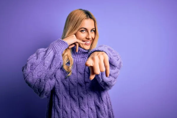 若い美しいブロンドの女性が紫色の背景にカジュアルなタートルネックセーターを着て 電話のジェスチャーで話しているとあなたを指して笑っています 電話してくれ — ストック写真