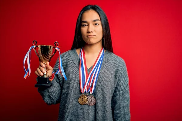 Young beautiful champion asian woman holding trophy wearing medals over red background with a confident expression on smart face thinking serious