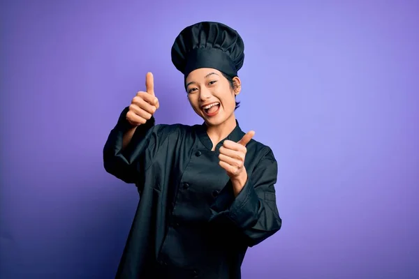 年轻美丽的中国厨师女士身穿炊具制服 头戴紫色背景的帽子 手握积极的手势 微笑着竖起大拇指 为成功感到高兴 赢的手势 — 图库照片