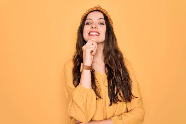 Young beautiful woman wearing casual t-shirt and diadem standing over yellow background looking confident at the camera with smile with crossed arms and hand raised on chin. Thinking positive.