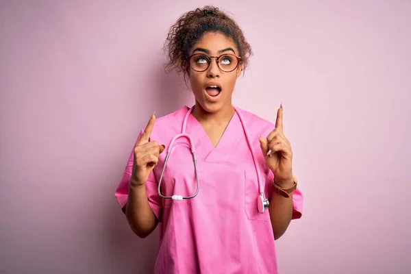 African american nurse girl wearing medical uniform and stethoscope over pink background amazed and surprised looking up and pointing with fingers and raised arms.