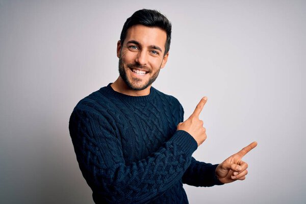 Young handsome man with beard wearing casual sweater standing over white background smiling and looking at the camera pointing with two hands and fingers to the side.