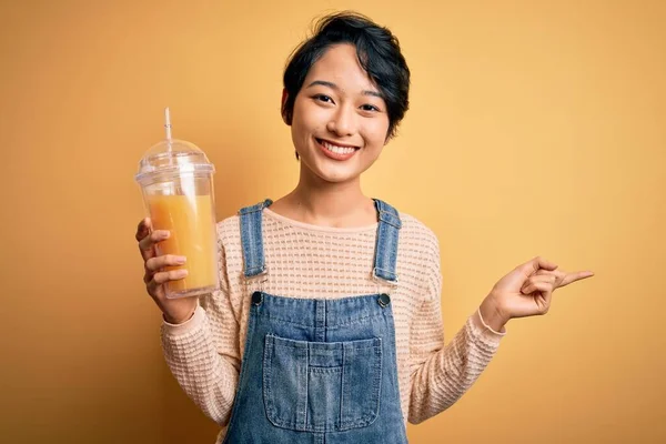 年轻美丽的中国女人 喝着健康的橙汁 背负着孤立的黄色背景 非常快乐 手指手画脚 — 图库照片