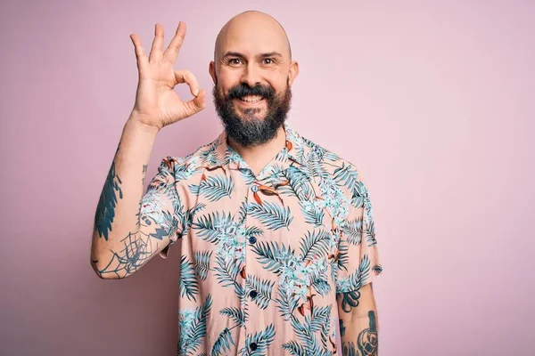 Sakallı, dövmeli, pembe arka planda sıradan bir çiçek gömleği giyen yakışıklı kel adam el ve parmaklarıyla tamam işareti yapıyor. Başarılı ifade.