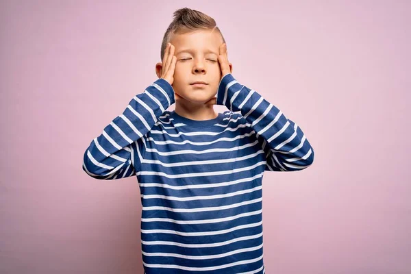 青い目の若い白人の子供は 痛みと片頭痛のために絶望的でストレスを受けたピンクの背景にネイビーのストライプのシャツを着ていました 手を頭に — ストック写真