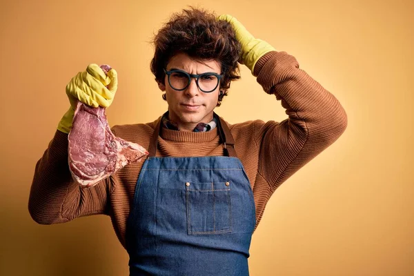 Young handsome butcher man holding meet steak standing over isolated yellow background confuse and wondering about question. Uncertain with doubt, thinking with hand on head. Pensive concept.