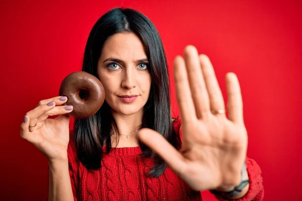 年轻的黑发女子 蓝眼睛 吃巧克力甜甜圈 背靠红隔离的背景 张开手 做停止手势 严肃而自信的表情 防守姿势 — 图库照片