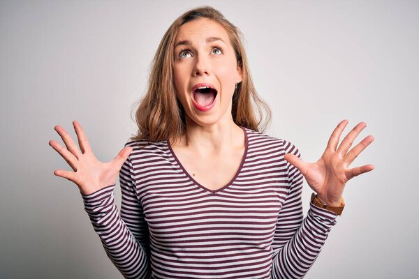 Молодая красивая блондинка в повседневной полосатой футболке на изолированном белом фоне празднует сумасшедший и сумасшедший от успеха с поднятыми руками и закрытыми глазами, крича взволнованно. Концепция победителя
