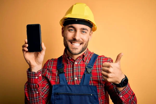 年轻的建筑工人戴着安全帽 带着智能手机屏幕在黄色的背景上 高兴地看到大大的笑容 做得很好 用手指竖起大拇指 很好的标志 — 图库照片