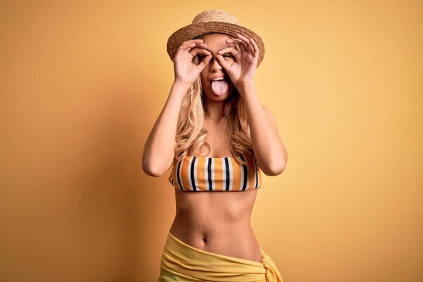 Young beautiful blonde woman on vacation wearing bikini and hat over yellow background doing ok gesture like binoculars sticking tongue out, eyes looking through fingers. Crazy expression.