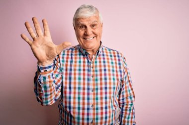 Üst düzey yakışıklı, parlak gömlekli, pembe arka planda yalnız gezen, kendinden emin ve mutlu bir şekilde gülümseyen, beş numarayı gösteren bir adam..