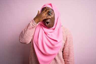 Genç Afro-Amerikan artı cüsseli kadın tecrit edilmiş pembe arka plan üzerine Müslüman başörtüsü takıyor. Şok olmuş bir şekilde yüzünü ve gözlerini eliyle örtüyor, utanmış bir ifadeyle parmaklarından bakıyor..