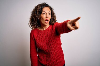 Orta yaşlı esmer kadın izole edilmiş beyaz arka planda dikilen sıradan bir süveter giyiyor. Parmağı önde, ağzı açık, şaşırmış bir ifadeyle işaret ediyor.