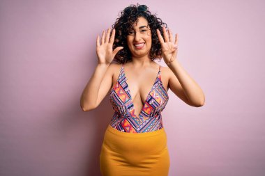 Genç, güzel bir Arap kadın, pembe arka planda mayo ve güneş gözlüğü takıp dokuz numarayı gösterirken kendine güvenen ve mutlu bir şekilde gülümsüyor..