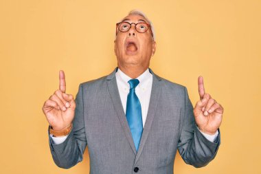 Orta yaşlı, kır saçlı, yakışıklı iş adamı sarı arka planda gözlük takıyor. Şaşırmış ve şaşırmış bir şekilde yukarı bakıyor, parmaklarıyla işaret ediyor ve kollarını kaldırıyor..