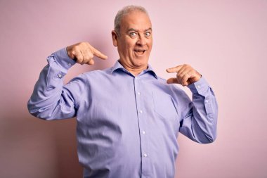 Orta yaşlı yakışıklı, beyaz gömlekli, pembe arka planda dikilmiş, neşeli gülümseyen, parmaklarıyla dişlerini ve ağzını gösteren bir adam. Diş sağlığı konsepti.