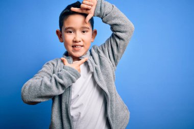 Küçük çocuk mavi izole edilmiş arka planda spor kazak giyiyor gülümsüyor ve elleri ve parmakları gülen yüzlü bir çerçeve yapıyor. Yaratıcılık ve fotoğrafçılık kavramı.