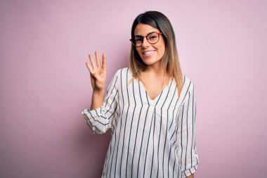 Genç ve güzel bir kadın pembe arka planda günlük çizgili tişört ve gözlük takıyor ve parmaklarıyla dört numarayı gösterirken kendinden emin ve mutlu gülümsüyor..