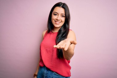 Young brunette woman wearing casual summer shirt over pink isolated background smiling cheerful offering palm hand giving assistance and acceptance. clipart