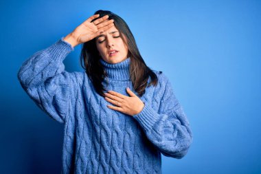 Mavi gözlü, balıkçı yaka kazak giyen genç esmer kadın hastalık, ateş, grip ve soğuk algınlığı için alnına dokunuyor.