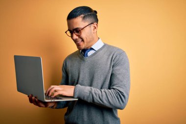 Genç Brezilyalı iş adamı dizüstü bilgisayarını kullanarak çalışıyor. Sarı arka planda duruyor. Mutlu bir yüzü var ve gülümsüyor.