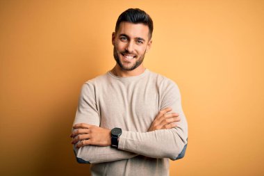 İzole edilmiş sarı arka planda dikilip kollarını kavuşturup kameraya bakarken gülümseyen genç, yakışıklı bir adam. Pozitif kişi.