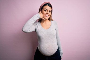 Genç, güzel, esmer bir kadın hamile, pembe arka planda bebek bekliyor. Gülümsüyor, el ve parmaklarıyla telefonla konuşuyor gibi. İletişim kavramları.