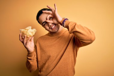 Genç Brezilyalı elinde patates kızartmasıyla izole edilmiş sarı arka planda gülümseyen ve el pençe divan duran bir adam.