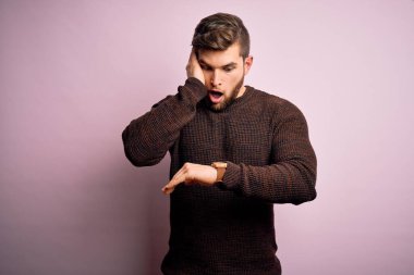 Sakallı ve mavi gözlü genç bir adam pembe arka planda sıradan bir süveter giyiyor. Endişeli saate bakıyor, geç kalmaktan korkuyor.