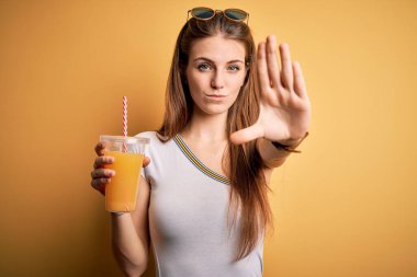 Genç ve güzel kızıl saçlı kadın sarı arka planda sağlıklı portakal suyu içiyor. Açık elle dur işareti yapıyor. Ciddi ve kendinden emin bir ifadeyle, savunma hareketi yapıyor.