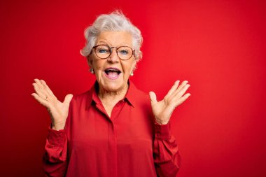 Son sınıf güzeli gri saçlı kadın kırmızı arka planda günlük gömlek ve gözlük takıyor çılgın ve hayret verici başarısını kutluyor kolları havada ve gözleri açık bir şekilde heyecanla haykırıyor. Kazanan konsept