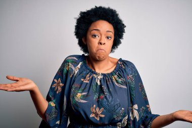 Genç, güzel, kıvırcık saçlı, çiçekli bir elbise giyen, elleri ve kolları havada, kafası karışık, Afro-Amerikan bir kadın. Şüphe kavramı.