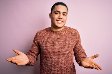 İzole edilmiş pembe arka planda duran sıradan bir süveter giyen genç Brezilyalı bir adam. Kolları ve elleri havada, kafası karışık bir ifade takınıyor. Şüphe kavramı.