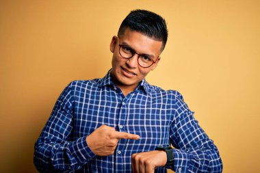 Sarı arka plan üzerine gündelik tişört ve gözlük takan genç, yakışıklı latin adam saati, sabırsızlığı ve gecikmeyi işaret ederek acele ediyor.