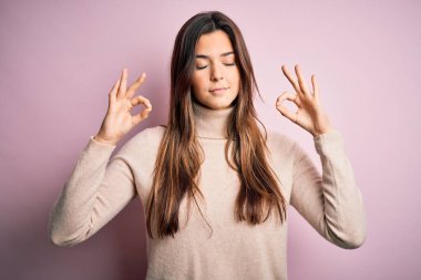Balıkçı yaka kazak giyen genç ve güzel bir kız izole edilmiş pembe arka planda dikiliyor rahatlıyor ve parmaklarıyla meditasyon hareketi yaparken gözleri kapalı gülümsüyor. Yoga konsepti.